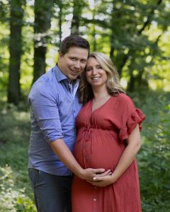Liebespaar hält den Babybauch fest und lacht in die Kamera.