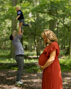 Im Vordergrund steht eine werdende Mutter und im Hintergrund wirft der Vater das Kind in die Luft.