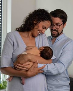 Die Mutter hält das Baby am Arm und die Eltern lachen es an.