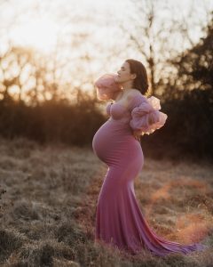 eine schwangere Frau steht mit einem glamourösen Kleid bei Sonnenuntergang auf einer Wiese.